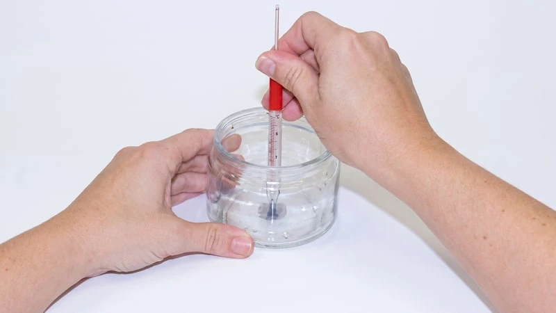 Hands centering a wick in an empty glass jar using a slim red thermometer