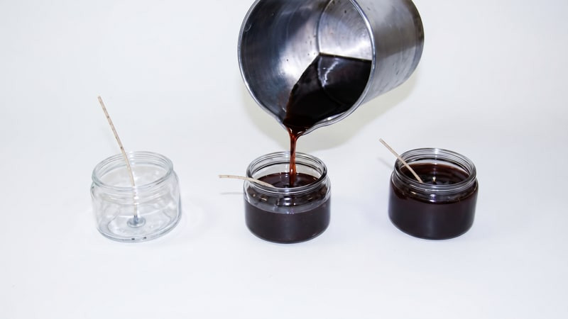 Dark red wax poured from a metal pitcher into a wicked glass jar, empty and filled jars alongside