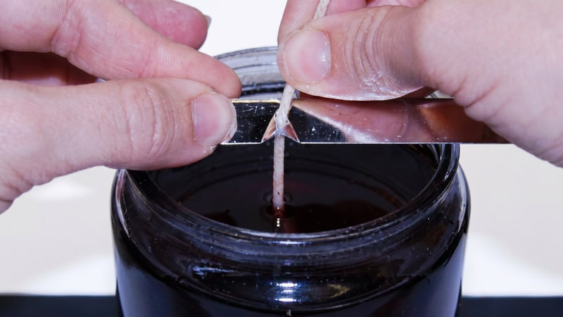 Hands holding a wick centered with a metal bar across a jar of dark red wax
