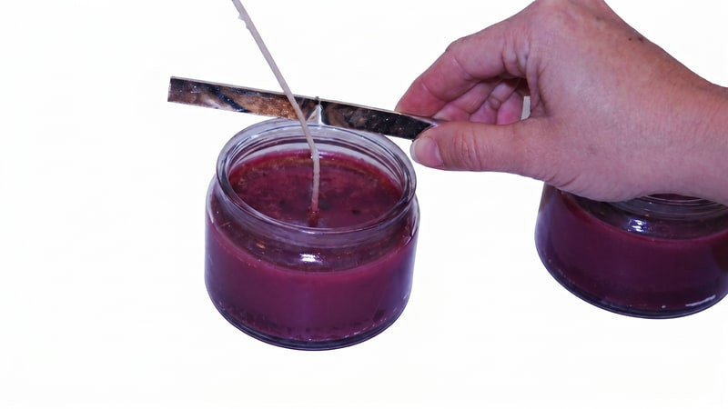 Hand setting a wick straight against a metal bar laid across a jar of poured purple-red wax