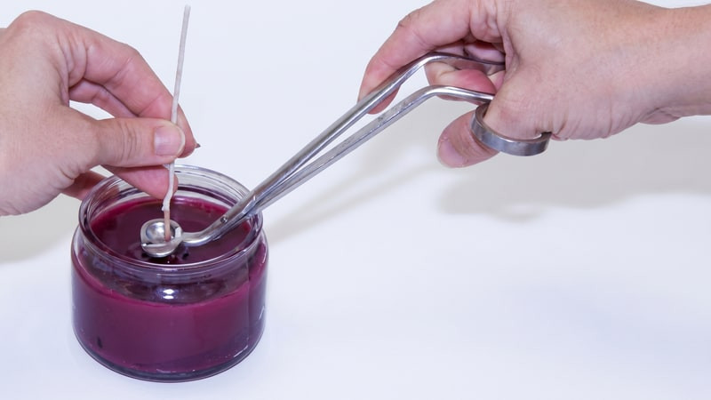 Hands ladling wax around the wick of a magenta container candle with a long metal spoon