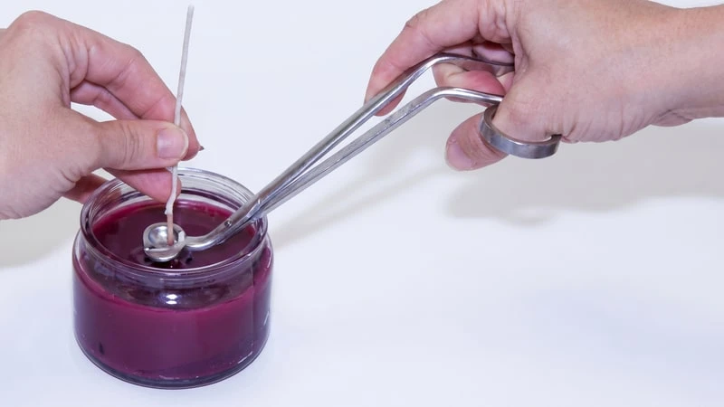 Hands ladling wax around the wick of a magenta container candle with a long metal spoon
