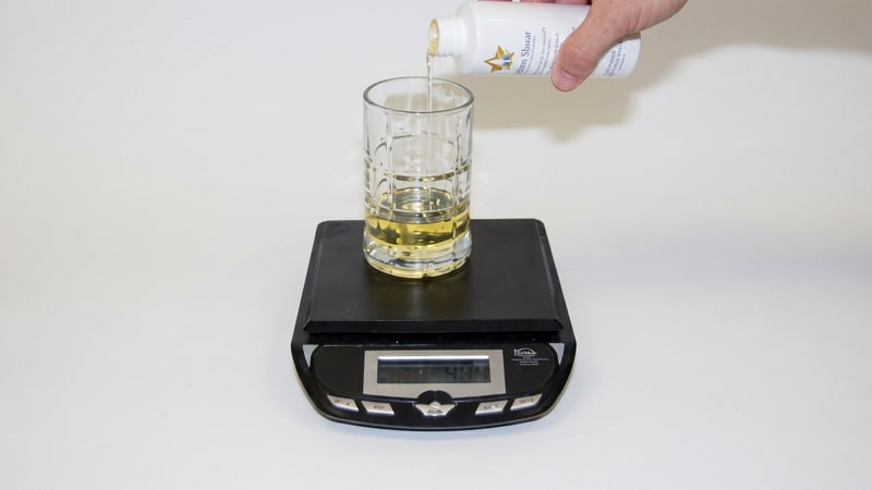 Hand pouring pale fragrance oil from a Lone Star Candle Supply bottle into a glass cup on a digital scale