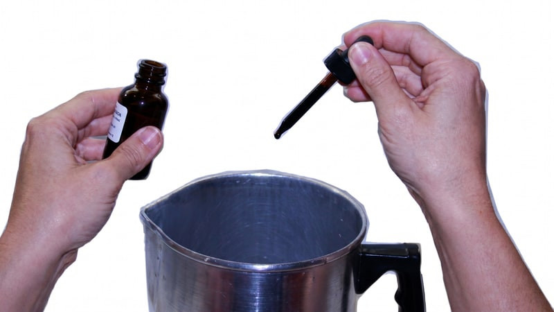 Hands holding an amber dropper bottle and dropper above an aluminum pouring pitcher