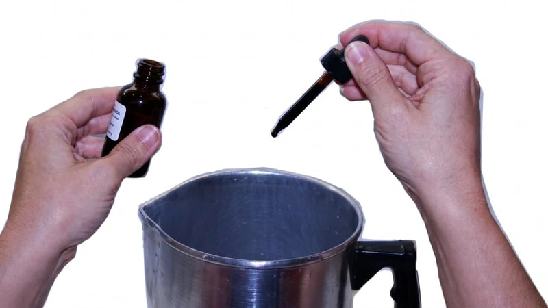 Hands holding an amber dropper bottle and dropper above an aluminum pouring pitcher