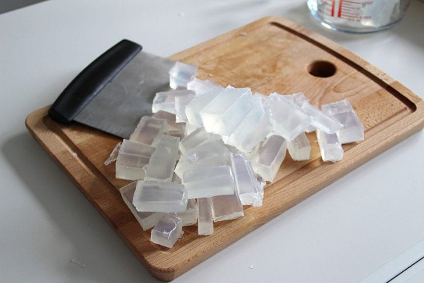 Chopped clear melt-and-pour soap base cubes on a wooden cutting board beside a black-handled bench scraper