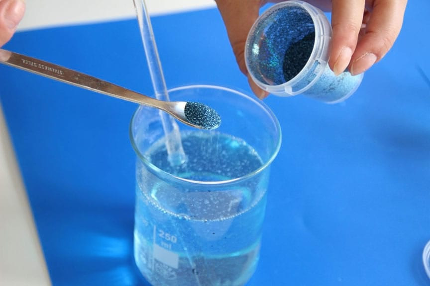 Spoon of blue glitter and a tipped tub held over a beaker of melted blue soap on a blue mat