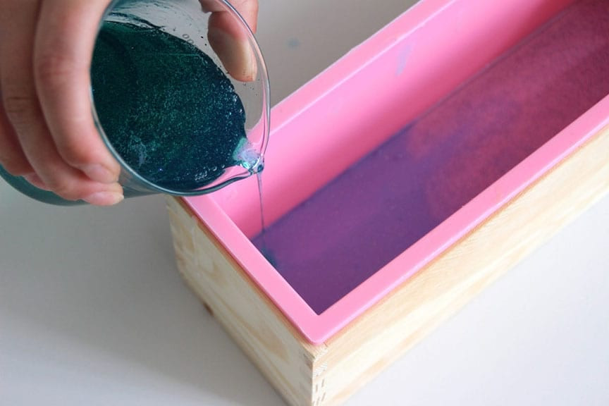 Teal glittered soap poured from a beaker into a pink silicone loaf mold over a purple layer