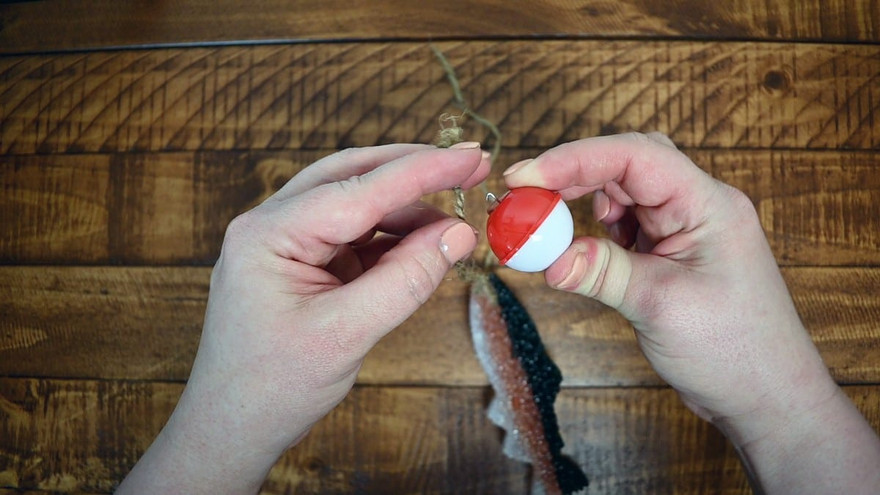 Hands attaching a red-and-white fishing bobber to twine on a baked trout-pattern freshie over wood