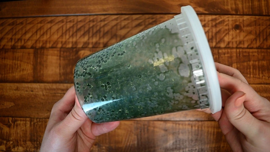 Hands shaking a sealed clear tub of green-tinted aroma beads, held sideways over a wood table