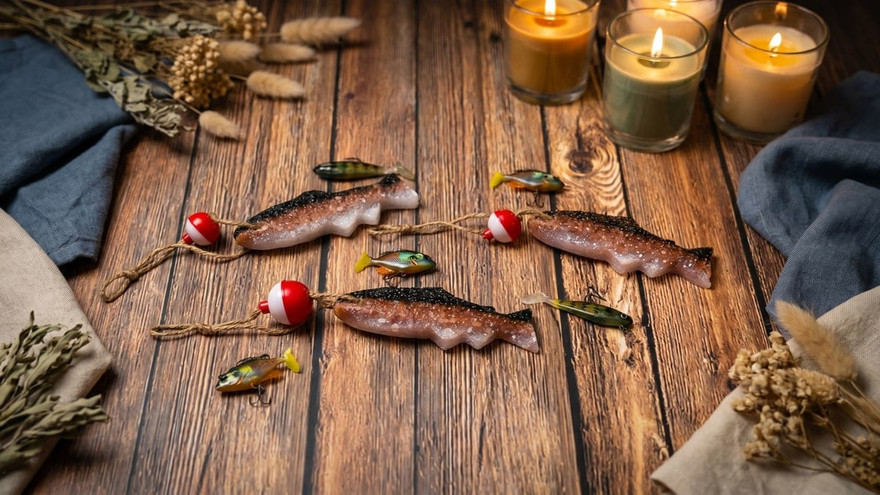 Three trout-shaped aroma-bead freshies with red-and-white bobbers and twine on weathered wood, lit candles and lures nearby