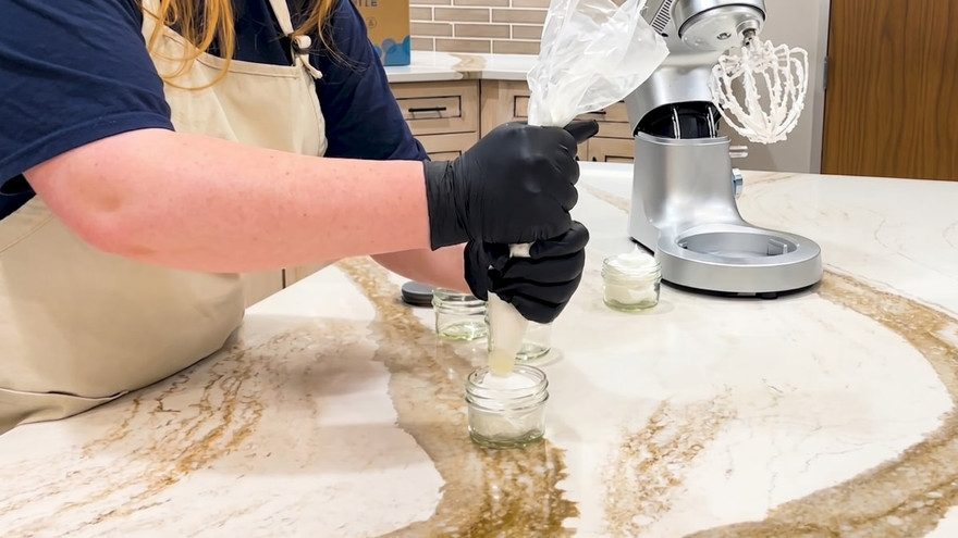 Black-gloved hands piping whipped white foaming bath butter scrub from a piping bag into a small glass jar on a marble counter, beside a stand mixer with a whisk attachment