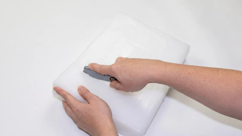 Hand scoring a white wax slab with a small gray hand tool on a white surface
