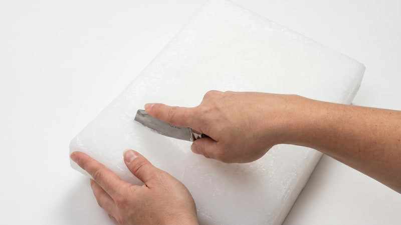Hand pressing a gray tool into a scored white wax slab to snap it on a white surface