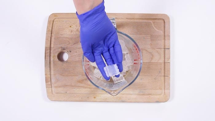 Blue-gloved hand placing white soap base cubes into a glass measuring jug on a wooden board