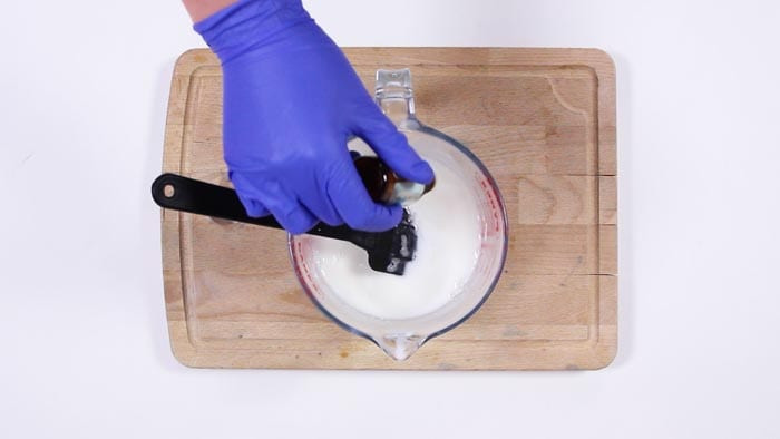 Blue-gloved hand adding fragrance oil from a black spoon into melted white soap
