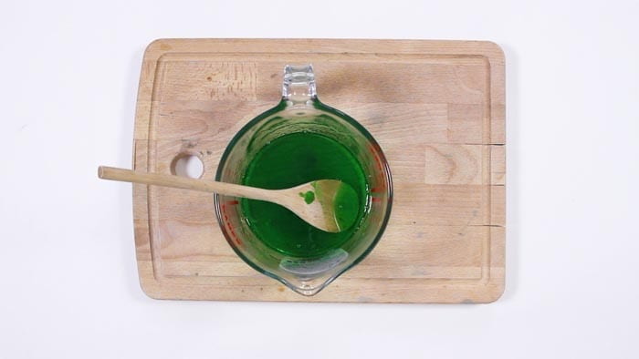 Glass measuring jug of bright green melted soap with a wooden spoon resting inside it