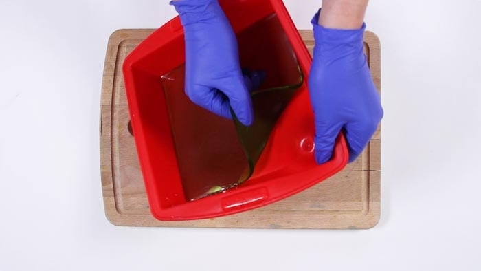 Blue-gloved hands flexing a red silicone mold to release a set sheet of green soap
