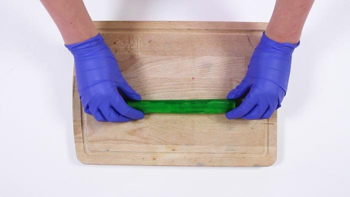 Blue-gloved hands rolling a thin green soap sheet into a log on a wooden board