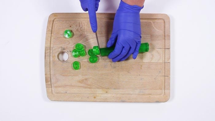 Blue-gloved hands slicing a rolled green soap log into spiral discs with a knife