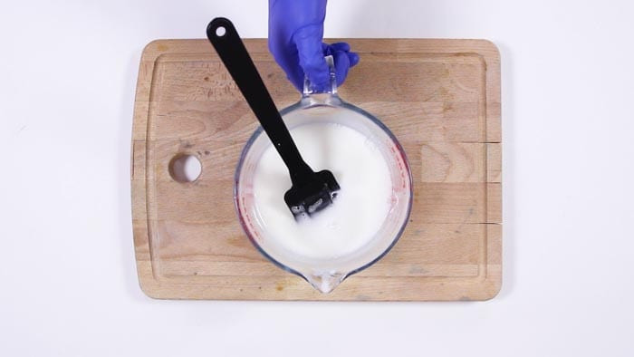 Blue-gloved hand stirring melted white soap with a black spatula in a glass jug
