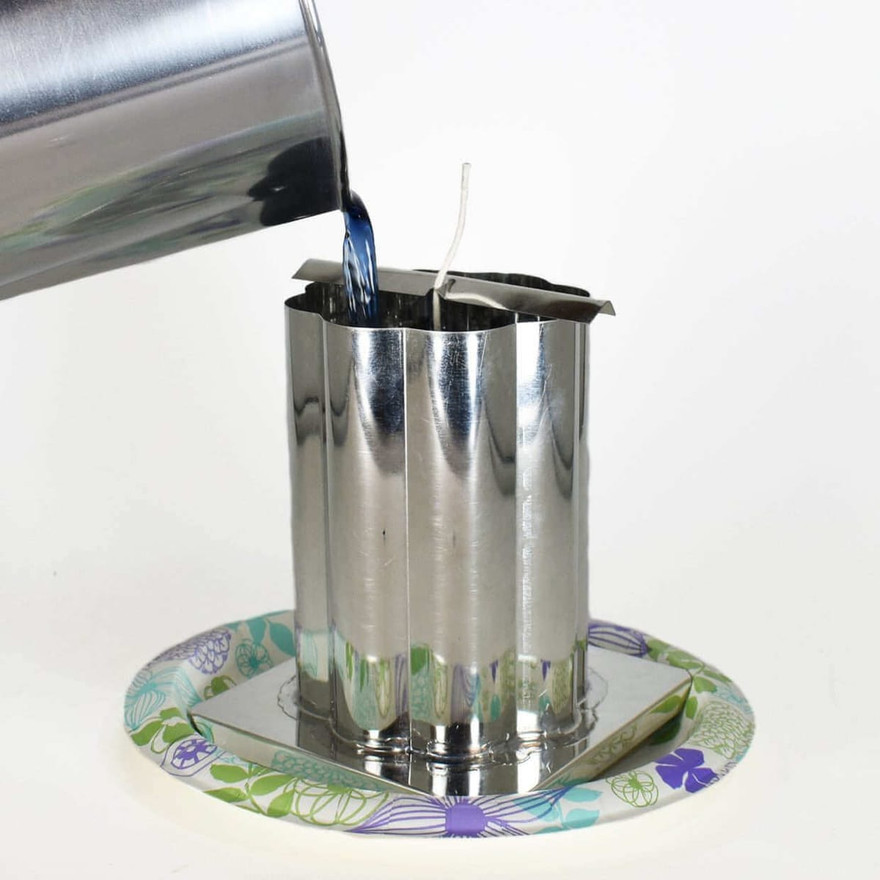 Blue melted wax poured from a steel pot into a wicked fluted pillar mold on a floral plate