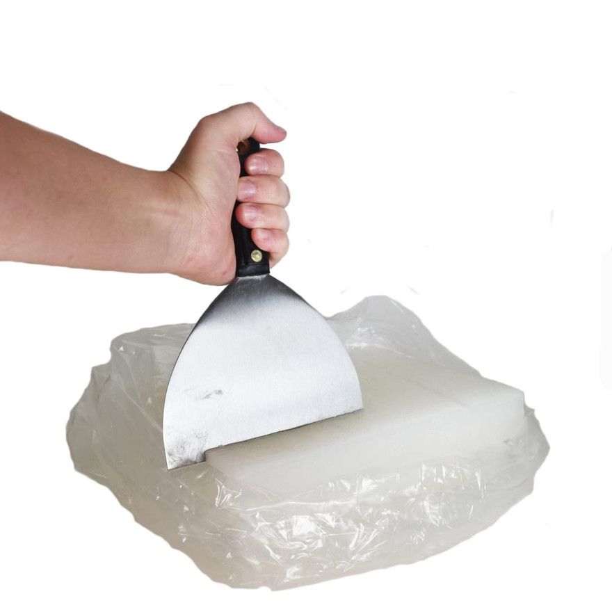 Hand pressing a putty knife into a white paraffin wax slab wrapped in plastic