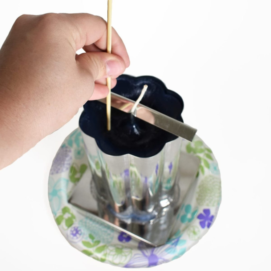 Hand poking a bamboo skewer into dark blue wax around the wick of a fluted pillar mold