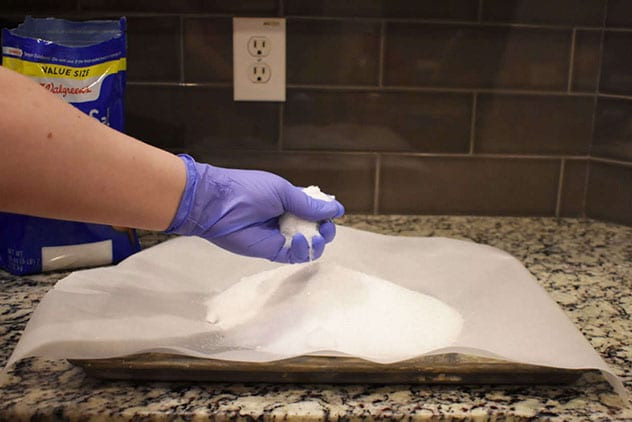 Purple-gloved hand crumbling a clump of Epsom salt over a parchment-lined baking sheet