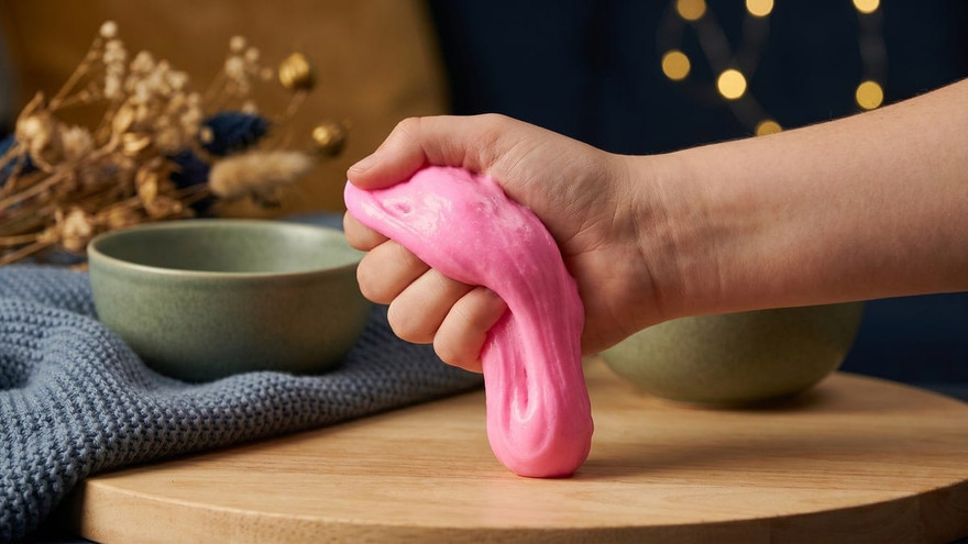 Hand squeezing a stretchy ball of bright pink slime on a wood board, cozy styled backdrop