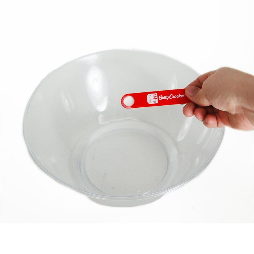 Hand holding a red Betty Crocker quarter-teaspoon measuring spoon over an empty clear plastic bowl