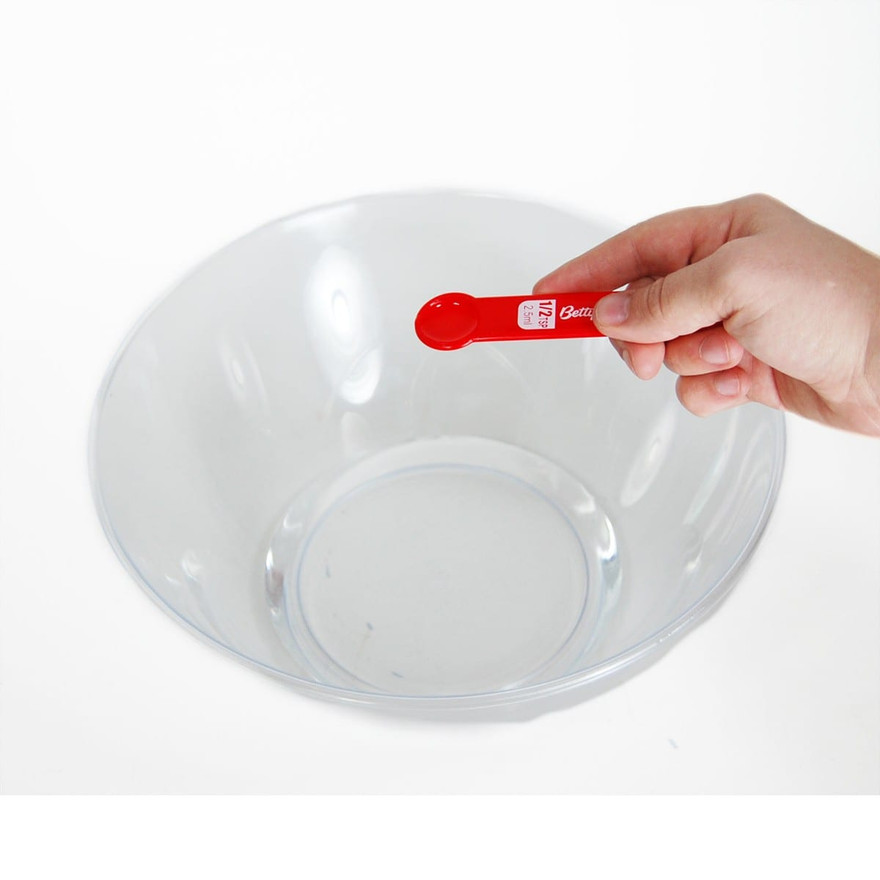 Hand holding a red half-teaspoon measuring spoon over an empty clear plastic bowl