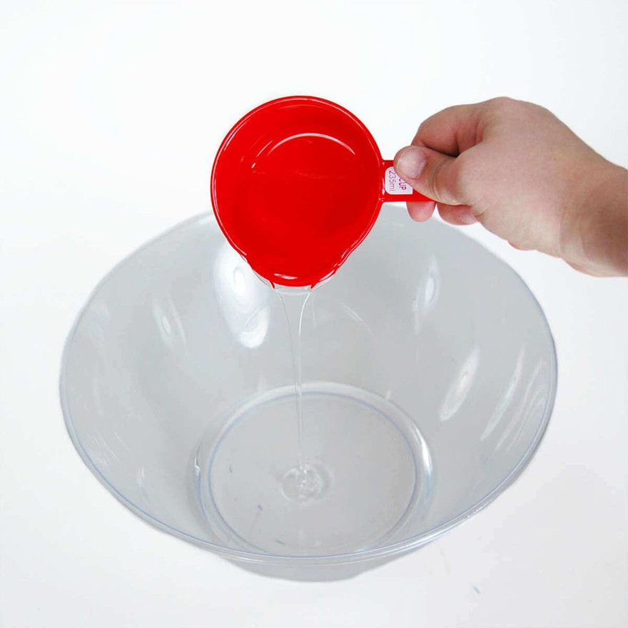 Hand pouring clear glue from a red measuring cup into a clear plastic mixing bowl