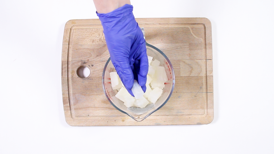 Purple-gloved hand placing white soap base cubes into a glass measuring bowl on a wooden cutting board