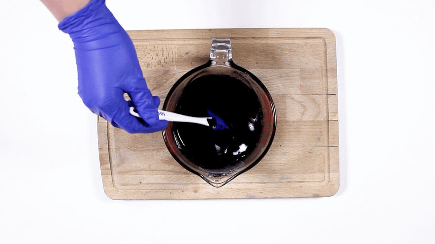 Blue-gloved hand stirring black-dyed melted soap in a glass measuring jug with a white spatula