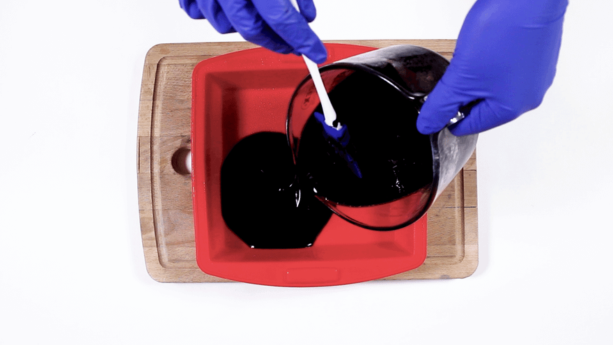 Blue-gloved hands pouring black melted soap from a glass jug into a red square silicone mold