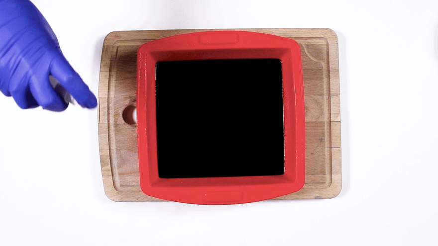 Red square silicone mold filled with smooth black soap, blue-gloved hand pointing nearby