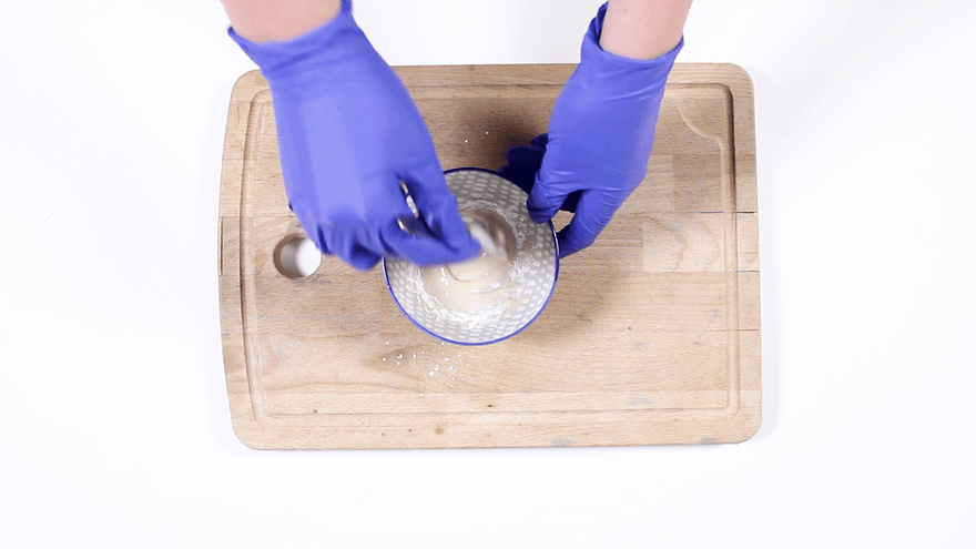 Blue-gloved hands mixing a creamy white paste in a small dotted bowl on a cutting board