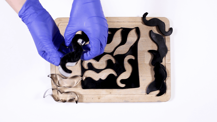 Blue-gloved hands cutting moustache shapes from black soap with metal cutters on a wooden board