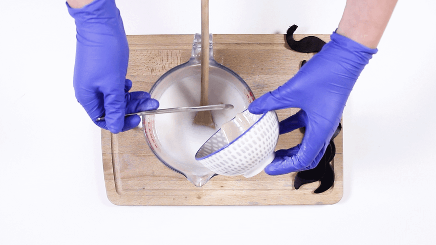 Blue-gloved hands straining white melted soap through a dotted bowl into a glass jug, black moustaches alongside