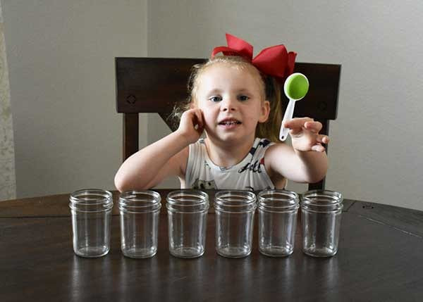 Young girl with red hair bow holding a green measuring spoon behind six empty glass jars on a table