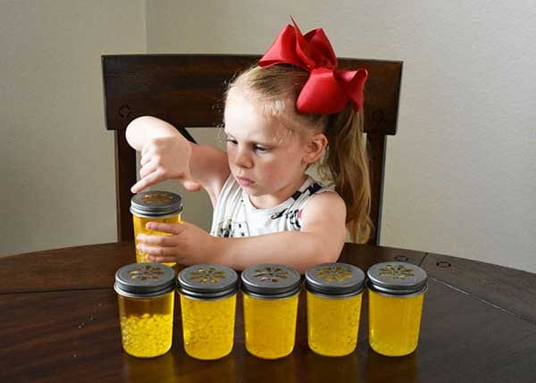 Girl pressing a daisy-cut lid onto a yellow gel jar beside five capped jars on a wood table