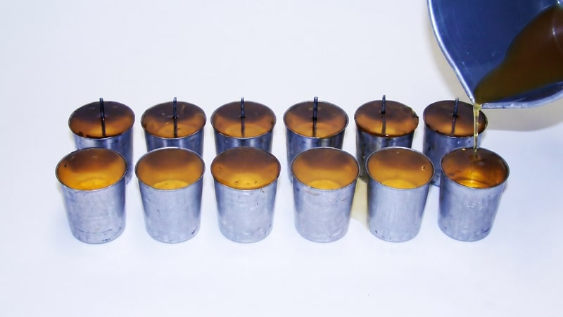Amber wax poured from a blue pitcher into two rows of metal votive molds with wick pins