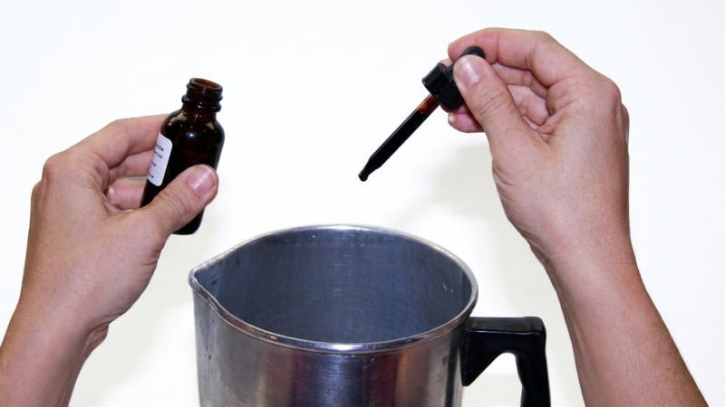 Hands holding an amber dye bottle and dropper over a steel pouring pot