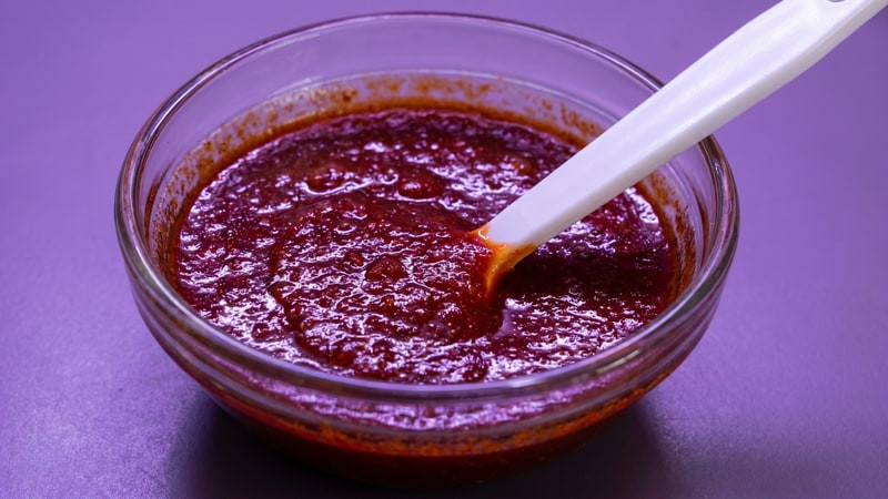 Reddish-brown thickened wax with a white spatula in a glass bowl on a purple surface