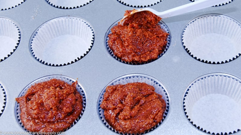 White spatula spooning reddish-brown whipped wax into cupcake liners in a baking tin