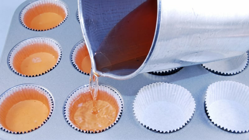Amber melted wax poured from a pitcher into orange-filled cupcake liners in a baking tin