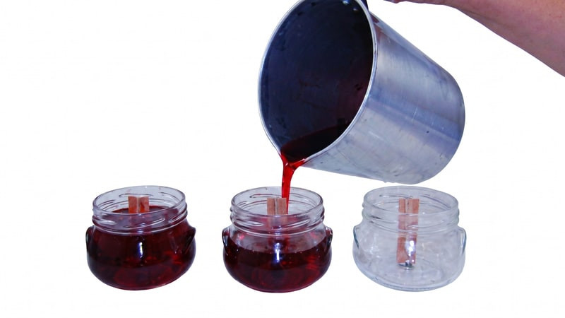 Dark red melted wax pouring from a steel pitcher into glass jars with wooden wicks