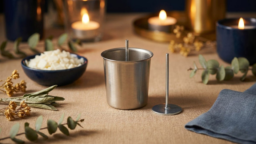 Metal votive mold with wick pin upright inside, a separate wick pin beside it, eucalyptus and tealights around