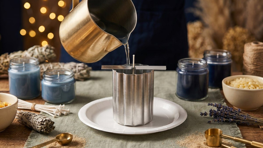 Melted wax pouring from a gold pitcher into a fluted metal mold on a paper plate, blue candles around
